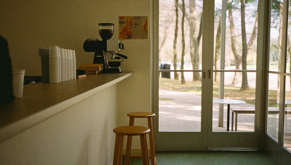 A small, plainly furnished cafe servery with a counter, coffee machine and glass doors looking out onto the park